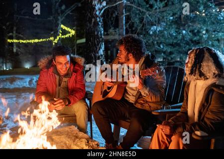 African young man playing guitar while sitting outdoors near the fire with his friends during winter holidays Stock Photo