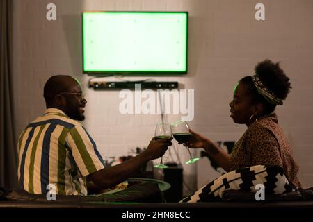 Happy African couple toasting with glasses of red wine and talking to each other while sitting on sofa and watching TV Stock Photo