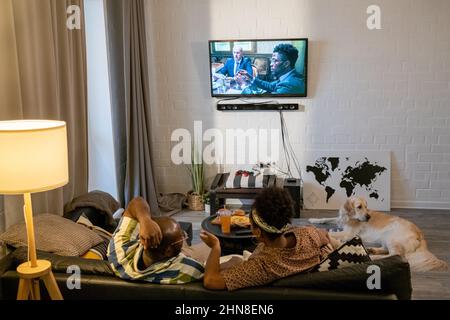 Rear view of African young couple sitting on sofa in front of the wall with television in the living room and discussing movie during watching Stock Photo