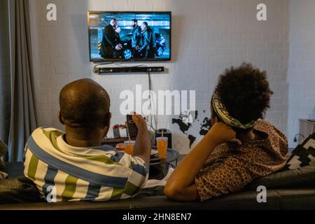 Rear view of African man using remote control to switch channels on tv hanging on the wall while sitting on sofa with woman Stock Photo