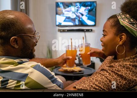Rear view of happy African couple toasting with glasses of juice and smiling to each other during watching the film at home Stock Photo
