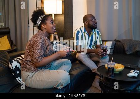 Excited African woman playing video game with her boyfriend watching for her game, they sitting on sofa in the living room Stock Photo