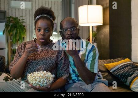 African young couple watching the movie with frightened look and eating popcorn together sitting on sofa in the living room Stock Photo