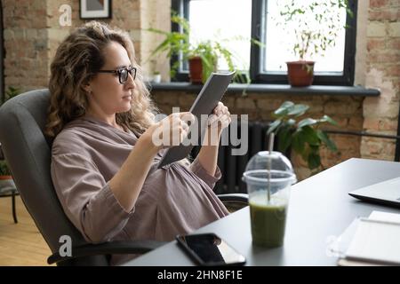 Pregnant businesswoman sitting on chair and reading documents in folder while sitting at the table with fresh cocktail Stock Photo
