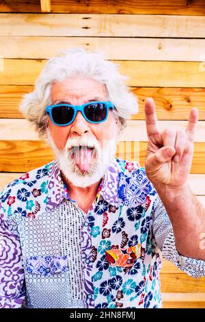 Vertical front portrait of old mature man with tongue and devil gesture. Fun senior people with colorful clothes and crazy expression. Alternative you Stock Photo