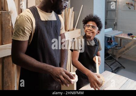 Portrait of smiling African-American boy looking at father while enjoying time together in carpentry workshop Stock Photo
