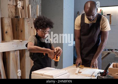 Side view portrait of cute African-American boy building wooden birdhouse and using power drill in workshop with father Stock Photo