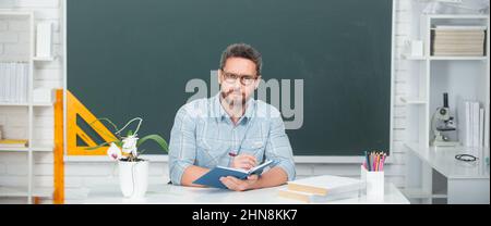 Young modern male teacher waiting for students. Teacher in casual clothes at blackboard with copy space in class at school. Stock Photo