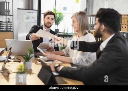 Multicultural business colleagues in stylish suits using modern gadgets and checking some documents during conference. International collaboration for common success. Stock Photo