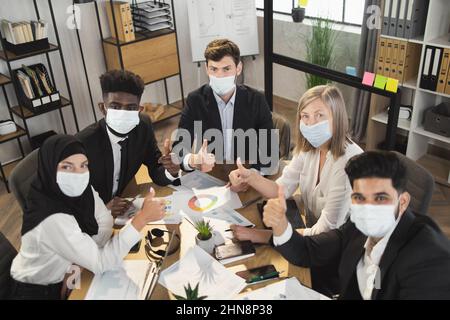 Five multi ethnic company workers in face masks and formal clothes showing thumb up while having meeting during pandemic. Business colleagues sitting at desk and looking at camera. Stock Photo