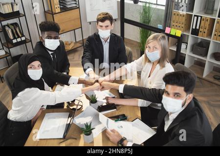 Five multi ethnic company workers in face masks and formal clothes fist bumping while having meeting during pandemic. Business colleagues sitting at desk and looking at camera. Stock Photo