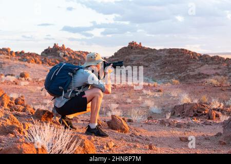 Photographer taking photo in rocks of Namib Desert, Namibia, Africa. Red mountains and sunset sky in background. Landscape photography Stock Photo
