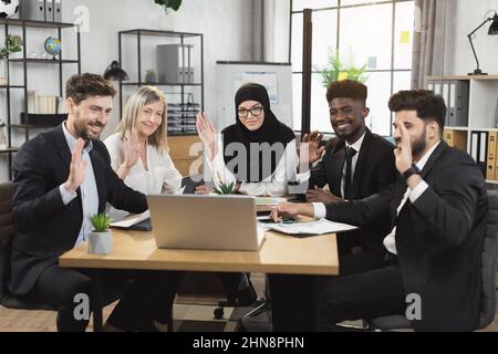 Group of multiracial company workers smiling and waving during conference call on modern laptop. Five business colleagues having online meeting at boardroom. Stock Photo