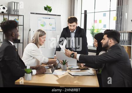 Group of busy multiracial people in formal clothes having active discuss during meeting at office. Men and women sitting at desk, using modern gadgets and examining various documents. Stock Photo