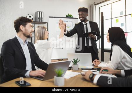 Positive african businessman giving high five to his multiracial colleagues during briefing at office. Successful negotiation of male and female partners. Stock Photo