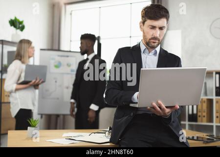 Caucasian man in suit using laptop and looking aside while leaning on desk at modern office. Multiracial company workers standing on background and talking. Stock Photo
