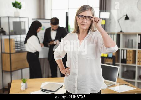 Portrait of confident mature woman in white shirt and eyewear posing at modern office. Blur background of two diverse coworkers talking near flip chart. Stock Photo