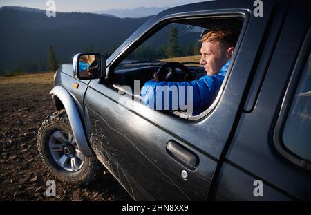 Close up view of young male blonde driving car, looking at landscape, riding on dirt roads in the mountains. Extreme off-road riding on mountain hills. Stock Photo