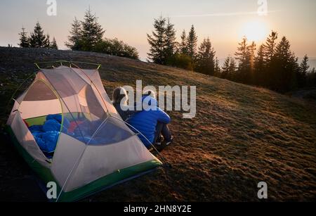 Back view of two travelers sitting on grass in front of tent set up on mountain hill, early in the morning and contemplating on sunrise. Soft sun rays lighting campsite. Stock Photo