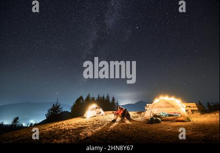 Girl hugging her boyfriend, they sitting together on grass between two tents iluminated by light bulbs and resting under beautiful starfull sky at night in the mountain hills. Stock Photo