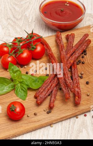Dried thin sausages on a wooden board with ketchup, cherry tomatoes and basil Stock Photo