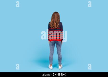 Full length back portrait of woman woman wearing striped casual style sweater, standing calm looking into the distance, waiting, posing backwards. Indoor studio shot isolated on blue background. Stock Photo