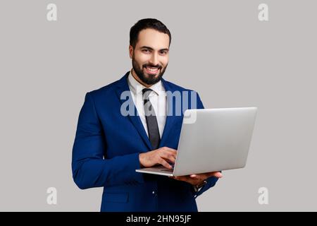 Positive bearded man holding laptop, looking at camera, surfing internet, freelancer working using computer, wearing official style suit. Indoor studio shot isolated on gray background. Stock Photo