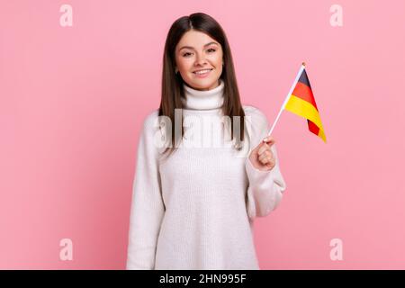 Satisfied positive female holding German flag in her hand and looking at camera with toothy smile, wearing white casual style sweater. Indoor studio shot isolated on pink background. Stock Photo
