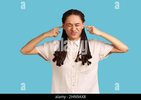 I dont want to listen it. Annoyed woman hears loud noise, avoids unpleasant sound, doesnt hear somebody's advice, closes ears with index fingers. Indoor studio shot isolated on blue background. Stock Photo