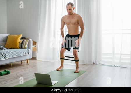 Fit sporty healthy man on a gym mat using rubber resistance exercise band doing legs stretching exercises, watching online training class on a laptop. Stock Photo