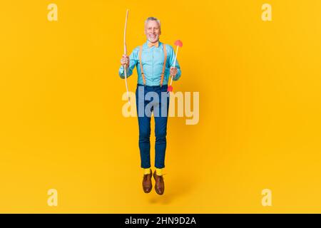 Photo of funky cheerful guy pensioner dressed teal shirt bowtie jumping shooting hearts isolated yellow color background Stock Photo