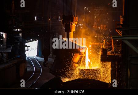 Molten metal pouring from metallurgy ladle. Workshop interior. Railroad and gate left. Stock Photo