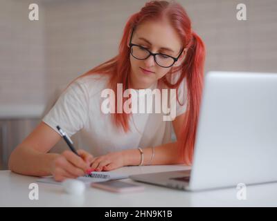 freelancer working on computer at home, making notes in notebook while listening to lecture or webinar. Attractive woman studying online using laptop software, web surfing or online shopping. Stock Photo