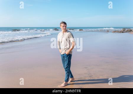 Attractive young caucasian man with full-arm tattoo in casual clothing walking on the beach barefoot. Scenic ocean view, morning. High quality photo Stock Photo
