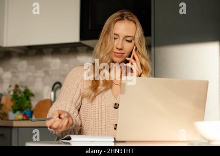 Young beautiful business woman with long fair hair in beige sweater talk on phone, make notes on notebook. Home office. Stock Photo