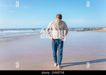 Attractive young caucasian man with full-arm tattoo in casual clothing walking on the beach barefoot. Scenic ocean view, morning. High quality photo Stock Photo