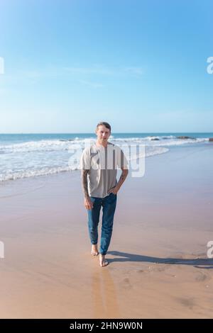 Attractive young caucasian man with full-arm tattoo in casual clothing walking on the beach barefoot. Scenic ocean view, morning. High quality photo Stock Photo