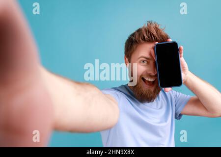 Portrait of positive optimistic bearded man blogger making selfie, point of view photo, covering eye with mobile phone, smiling happily. Indoor studio shot isolated on blue background. Stock Photo