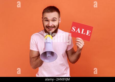 Portrait of smiling bearded man paying attention announcing big sale, holding loudspeaker, black Friday shopping, wearing pink T-shirt. Indoor studio shot isolated on orange background. Stock Photo