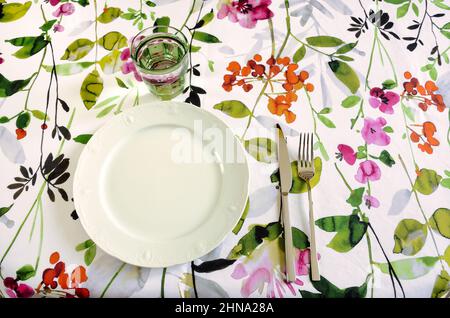 table set with floral tablecloth, white plate, green glass and stainless steel cutlery Stock Photo