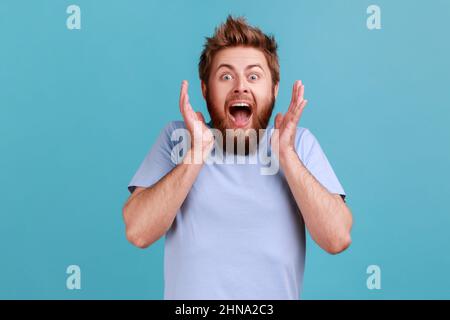 Portrait of pleasantly surprised glad bearded man smiles broadly shocked to receive unexpected news, looking at camera, expressing excitement. Indoor studio shot isolated on blue background. Stock Photo