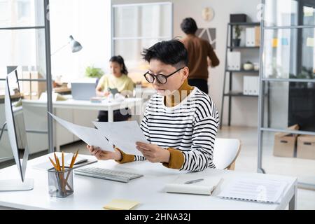 Asian young businesswoman in eyeglasses sitting at her workplace at office and examining the documents, she reading a contract Stock Photo