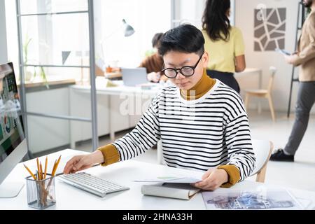 Asian serious businesswoman in eyeglasses sitting at the table and typing document on computer at office with her colleagues in the background Stock Photo