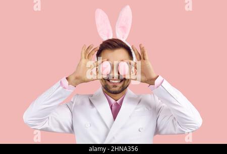 Happy young man in funny bunny ears smiling and covering his eyes with two Easter eggs Stock Photo