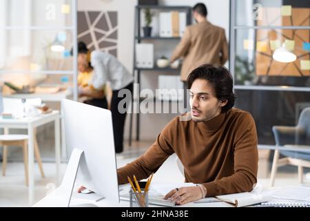 Young businessman sitting at the table at office and looking at computer monitor, he concentrating on his online work Stock Photo
