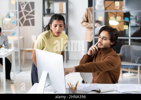 Young businessman pointing at computer monitor during his conversation on mobile phone, he showing online presentation to his colleague on computer mo Stock Photo
