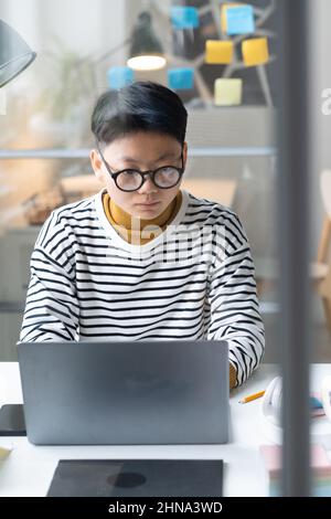 Serious Asian businesswoman in eyeglasses concentrating on her online work on laptop at the workplace at office Stock Photo