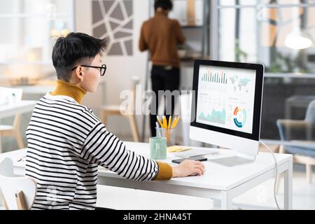 Rear view of businesswoman with short hair in eyeglasses sitting at the table in front of computer and analyzing the graphs on the monitor Stock Photo