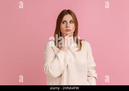 Portrait of blond woman standing deep in thoughts with puzzled serious doubtful face expression, making difficult decision, wearing white sweater. Indoor studio shot isolated on pink background. Stock Photo