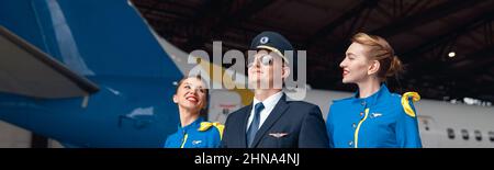 Happy pilot in uniform and aviator sunglasses walking together with two air stewardesses in blue uniform in front of big passenger airplane in airport Stock Photo